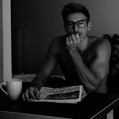 a shirtless man sitting at a table next to a coffee cup and reading a newspaper