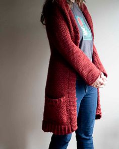 a woman standing in front of a white wall wearing a red cardigan sweater and jeans