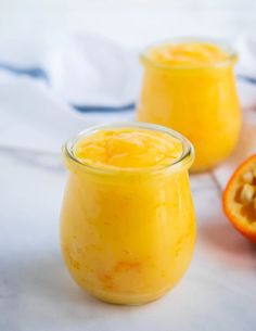 two jars filled with orange curd next to half an orange on a white surface