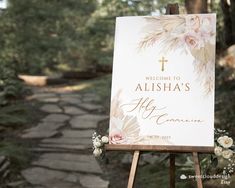 a welcome sign with flowers on it in front of a stone path and trees at an outdoor wedding venue