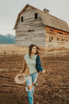 a woman walking across a dirt field with a hat in her hand and a barn in the background