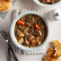 two bowls filled with meat and vegetables on top of a white table next to bread