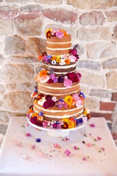 a multi layer cake with flowers on top sitting on a table next to a brick wall