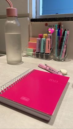 a pink notebook sitting on top of a table next to a bottle and pencils