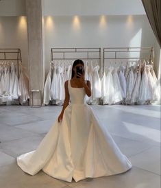 a woman in a white wedding dress taking a selfie with her cell phone while standing in front of a row of dresses