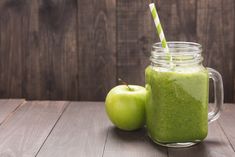 two green smoothies in mason jars next to an apple on a wooden table with a striped straw