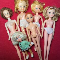 several dolls are posed next to each other on a red cloth covered surface, one is holding a green teddy bear and the other has blonde hair