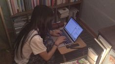 a woman sitting in front of a laptop computer on top of a wooden desk next to a bookshelf