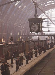 many people are walking on the platform near a train station with a clock hanging from the ceiling