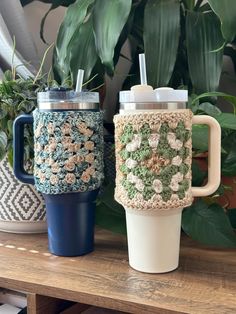 two crocheted coffee cups sitting on top of a wooden table next to potted plants