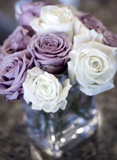 purple and white roses are in a clear vase on a counter top with other flowers