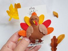 a hand holding a small felt turkey on top of a white table with fall leaves