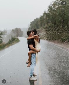 a man and woman kissing on the side of a wet road with trees in the background