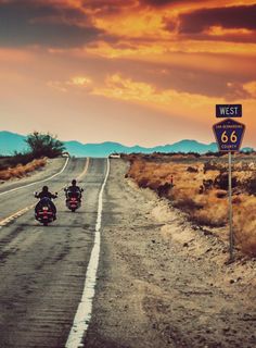 two people riding motorcycles down the road in front of a highway sign and sunset sky