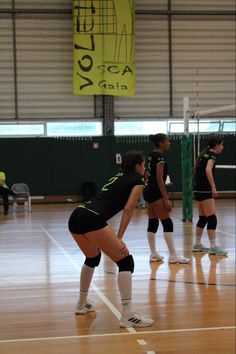 three women in black and green uniforms playing volleyball on an indoor court with people watching from the sidelines