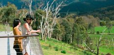 two women standing next to each other on a balcony overlooking a valley with trees and grass