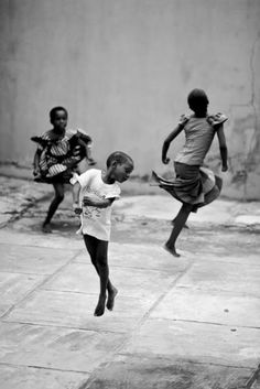 three young children are playing soccer on the sidewalk in front of a wall and building