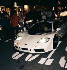 a white sports car driving down a street next to people standing on the side walk