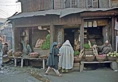 an old photo of people walking around in front of a building with vegetables on display