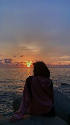 a woman sitting on top of a rock next to the ocean at sunset or sunrise