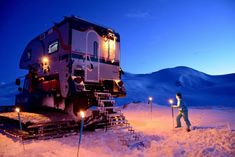 a man standing in the snow next to a large train car with lights on it