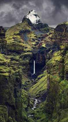 a waterfall in the middle of a green valley with snow capped mountain behind it and dark clouds overhead