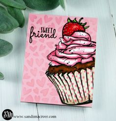 a card with a pink frosted cupcake and strawberry on it, sitting next to a plant