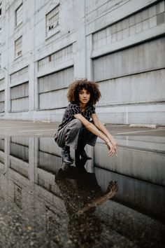 a young woman sitting on the edge of a puddle in front of a tall building