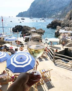 a person holding up a glass of wine on the beach with umbrellas and boats in the water