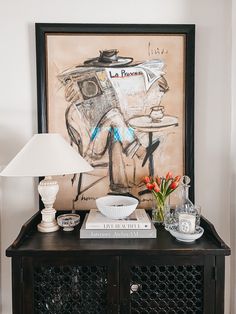 a painting on the wall above a table with books, vases and other items