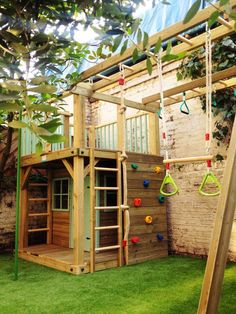 a wooden play structure with swings and climbing bars in the grass next to a brick wall