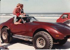 two people sitting on top of a red sports car at the beach with another person standing next to it