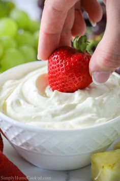 a hand dipping a strawberry into a bowl of cream cheese with grapes in the background