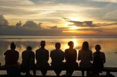 four people are sitting on a bench watching the sun go down over the water in front of them