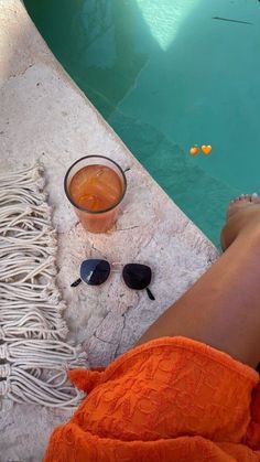 a person sitting on a towel next to a pool with sunglasses and a drink in it