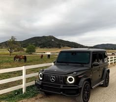 a black mercedes benz is parked in front of a white fence with horses grazing on the field behind it