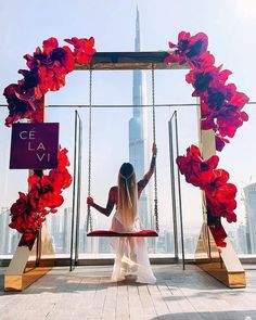 a woman sitting on a swing in front of a cityscape with red flowers