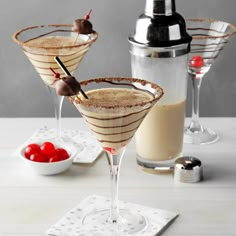 two martinis with chocolate and cherry garnish on the rim, next to a cocktail shaker