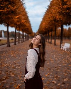 a woman standing in the middle of a park with trees lining both sides and leaves on the ground