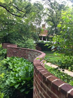 a brick wall in the middle of a garden with green plants and trees around it