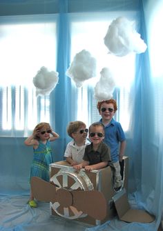 three children are posing for a photo in a cardboard box with clouds hanging from the ceiling