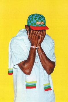 a man with his hands covering his face in front of a yellow background wearing a green checkered hat