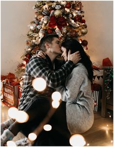 a man and woman kissing in front of a christmas tree with lights on the floor