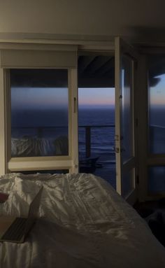 a laptop computer sitting on top of a bed next to two large windows overlooking the ocean