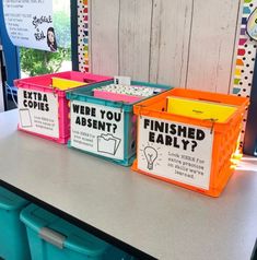 three plastic bins with labels on them sitting on a table in front of a bulletin board