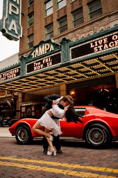 a man and woman kissing in front of a red car on the street with marquee lights behind them