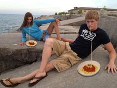 two people sitting on the rocks with plates of food in front of them and one person holding a fork