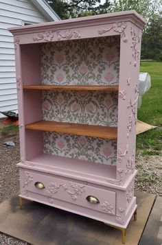 an old dresser painted pink and gold with floral wallpaper on the back, it is sitting outside