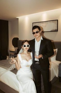 a man and woman standing next to each other in front of a bed holding wine glasses