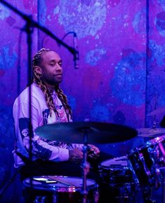 a man with dreadlocks playing drums in front of a purple and blue background